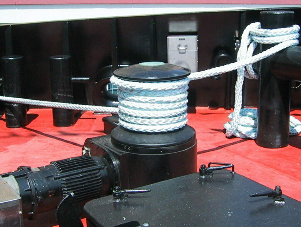 A close-up view of a ship's deck featuring a capstan wrapped with a thick  white rope. The capstan is mounted on a black base, with a motor visible on the side. Several black bollards can be seen in the background on the red deck, and the ship's side is visible in the upper part of the image. The deck equipment looks well-maintained and ready for use.