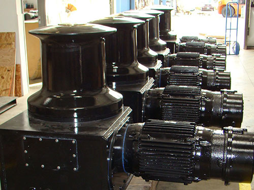 A row of newly manufactured capstans by Markey Machinery, arranged in a line on a workshop floor. The capstans are painted black and feature robust cylindrical bodies with flanged bases and integrated gear mechanisms, reflecting the industrial nature of the equipment.