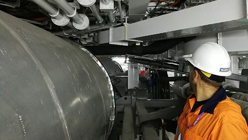 The image depicts an industrial setting where a person in an orange high-visibility jacket and a white safety helmet is inspecting large cylindrical machinery, potentially a winch or marine deck equipment.