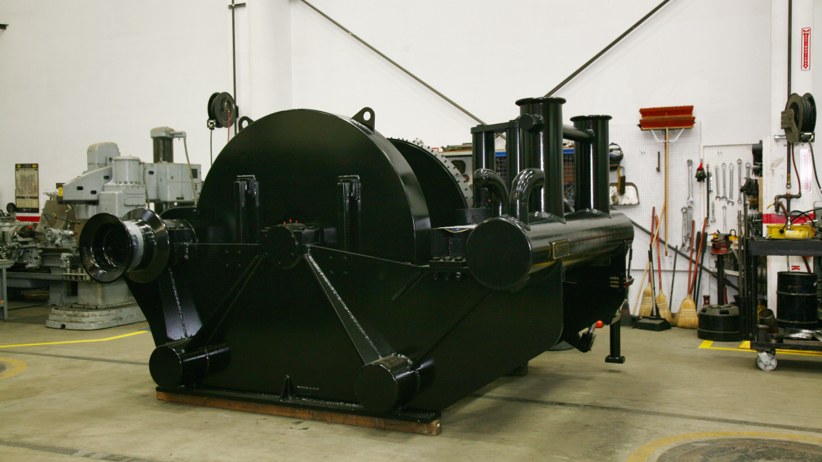 large, industrial winch painted black, situated in a workshop with various tools and equipment visible in the background. The winch is robust and appears to be designed for heavy-duty use, likely intended for installation on a tugboat as indicated by its design and context