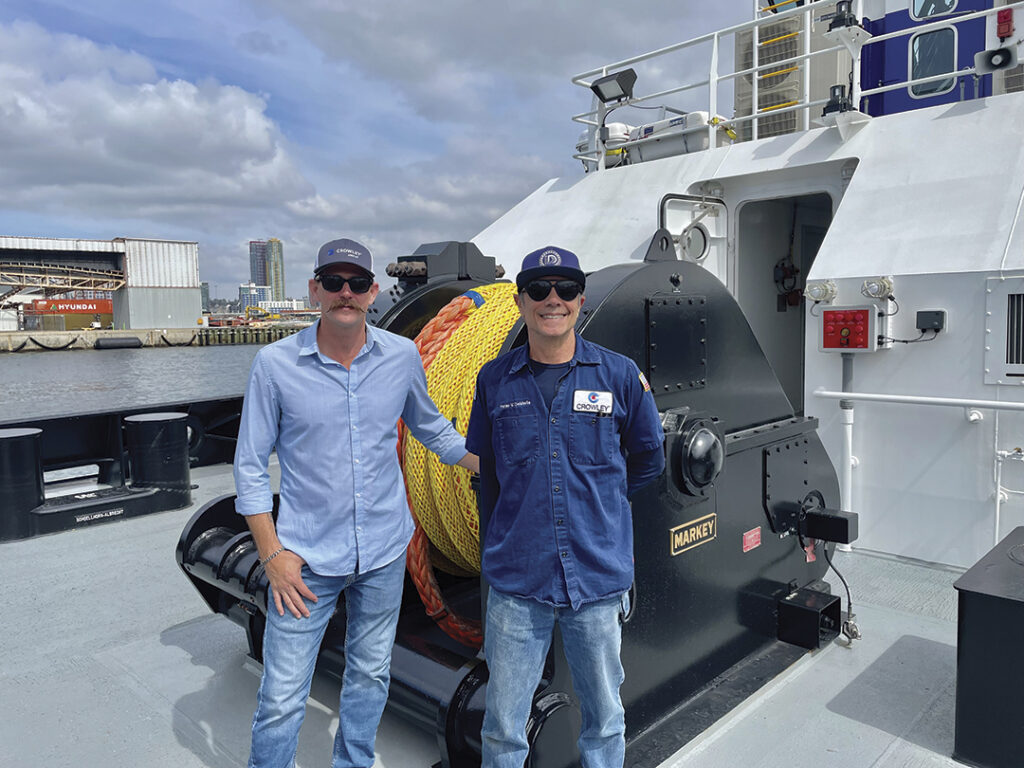 
Two men posing on the deck of the eWolf tugboat, with one wearing a light blue shirt and the other in a blue work uniform and cap. Behind them is a large industrial winch with a yellow rope. The background shows an industrial waterfront area with buildings and cranes under a partly cloudy sky. 