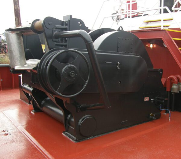 black winch mounted on a red deck. The winch has a prominent drum or spool with a thick chain wrapped around it. Several hand wheels and levers are visible on the winch, suggesting manual operation. There is a cylindrical object in the foreground, possibly a fairlead or roller to guide the chain. The winch appears to be well-maintained and in good operating condition.