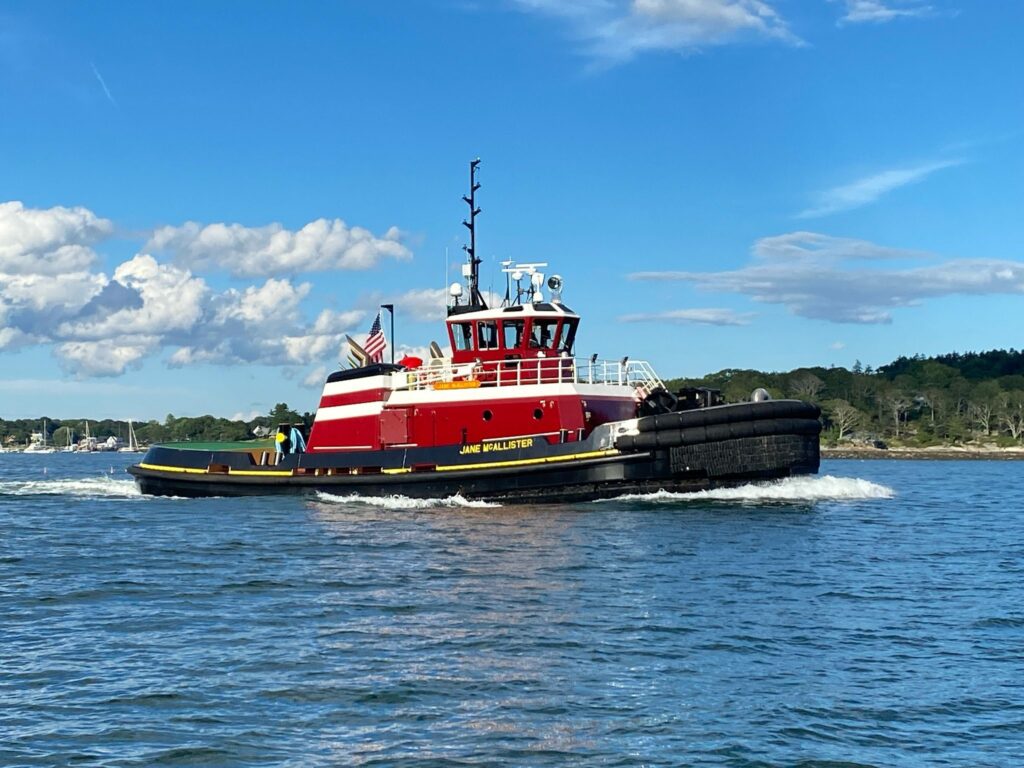 Red and white tugboat named "Jane McAllister" moving through a calm body of water under a bright blue sky with scattered clouds. The tugboat has an American flag displayed on the stern, and the shoreline with trees is visible in the background, indicating a coastal or river environment. The scene captures the tugboat in action, creating a small wake as it navigates through the water.