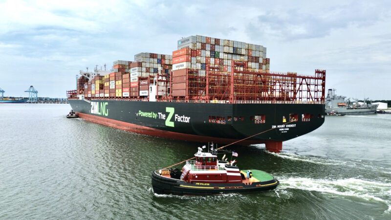 The image showcases a large container ship named "ZIM ENGLAND" in a harbor or port. The ship is predominantly white with black and green accents, and it's loaded with numerous colorful shipping containers stacked high. A smaller, black and red tugboat named "JANE MCALLISTER" is positioned alongside the ship, seemingly assisting with its maneuvering. The water is calm, reflecting the overcast sky. The background shows other vessels and port infrastructure in the distance.