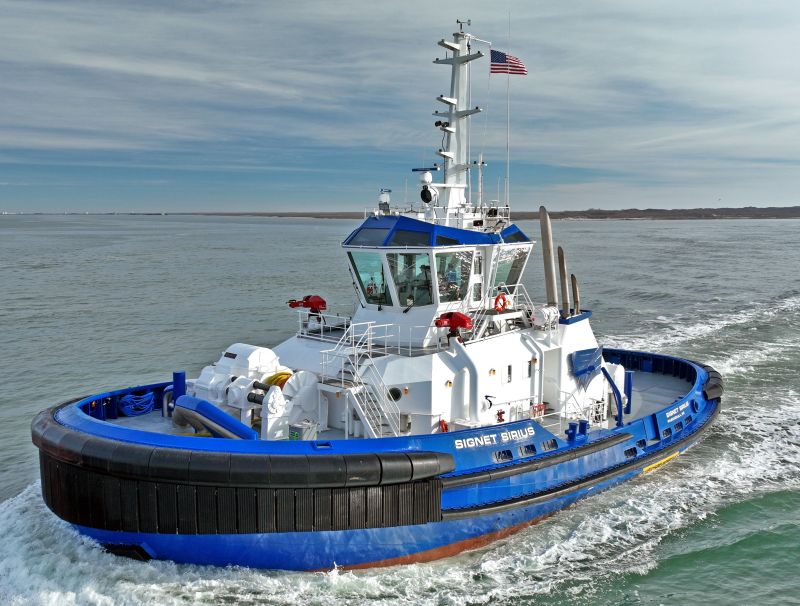 modern tugboat named Signet Sirius sailing on calm waters. The vessel has a blue and white exterior with black rubber bumpers on the bow for protection during towing operations. The tugboat is equipped with a sturdy bridge deck and multiple antennas, and an American flag flies from the mast. It appears to be moving at a steady pace with a clear sky and distant shoreline in the background.