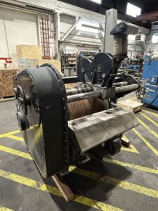 Markey Machine. large industrial winch inside a workshop or manufacturing facility. The winch has a heavy-duty spool wrapped with thick cable, likely used for lifting or pulling heavy loads. The machine appears to be mounted on wood blocks for stability, with various mechanical components visible, such as pulleys and protective covers. The surrounding area includes yellow safety markings on the floor and some industrial equipment and crates in the background.