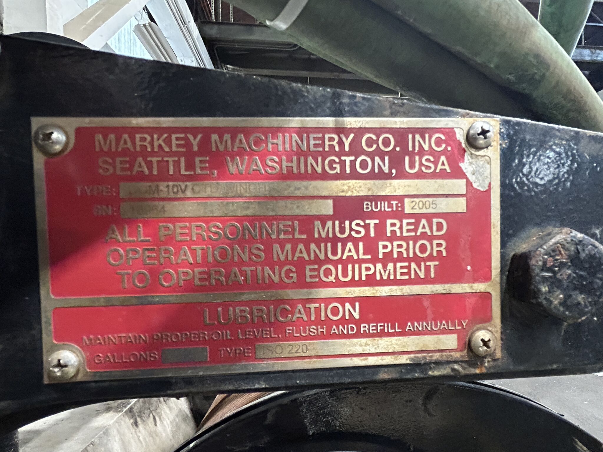 metal nameplate attached to machinery, reading: "Markey Machinery Co. Inc., Seattle, Washington, USA." It includes details like the model "Type: DYM-10V Cutting Winch" and "Built: 2005." There is a notice that states, "All personnel must read operations manual prior to operating equipment." At the bottom, it provides lubrication instructions, specifying to "Maintain proper oil level, flush and refill annually" with "ISO 220" oil type.