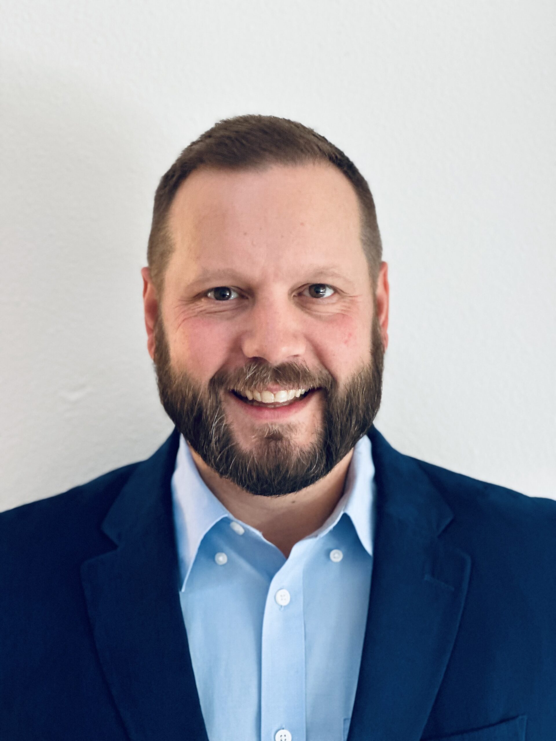 Peter Roney man with a friendly smile, a neatly trimmed beard, and short hair, dressed in a light blue button-down shirt and a navy blazer. He stands against a plain white background, giving the photo a clean and professional look.