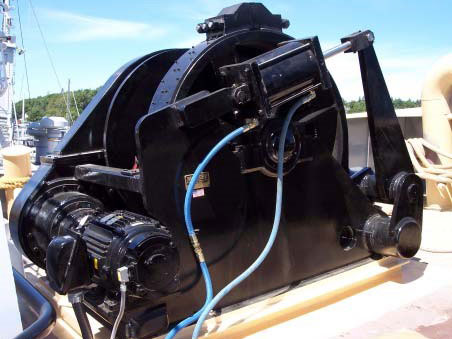 Markey Machine black industrial hawser winch system, likely designed for maritime use. It features robust components, including motorized mechanisms and hydraulic lines, mounted on a sturdy base with a backdrop of a sunny outdoor setting. The equipment is tailored for heavy-duty tasks such as towing or anchoring operations. Let me know if you'd like a specific focus or more detail in the description!