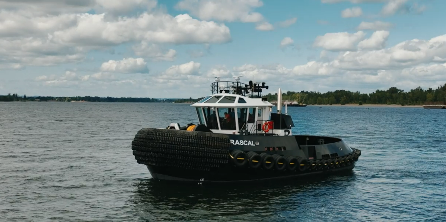 Hawser Winch black tugboat named "Rascal" floating on calm waters under a partly cloudy sky. The vessel is outfitted with large tire fenders along its sides for protection and features a white cabin structure with visible navigation equipment on top. It is positioned near a distant shoreline, suggesting a coastal or riverine setting.